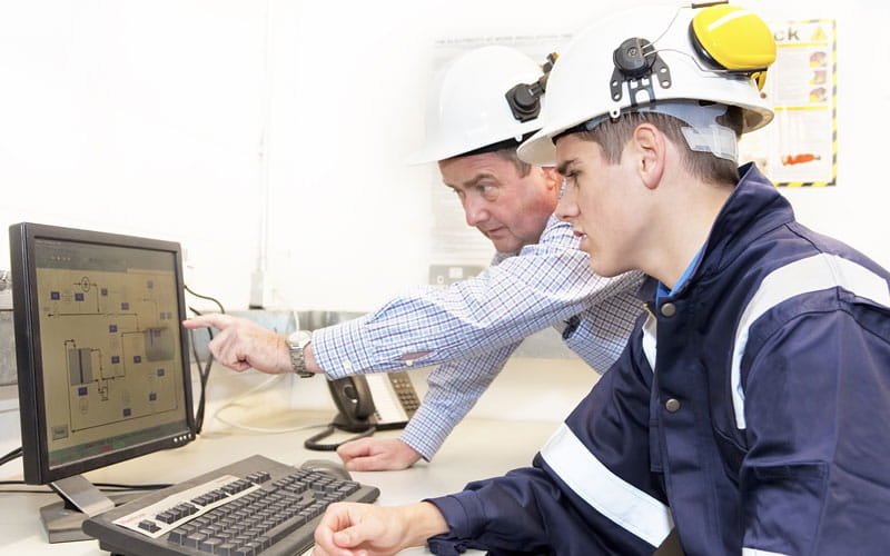 Two light-skinned men in hard hats work together on a computer