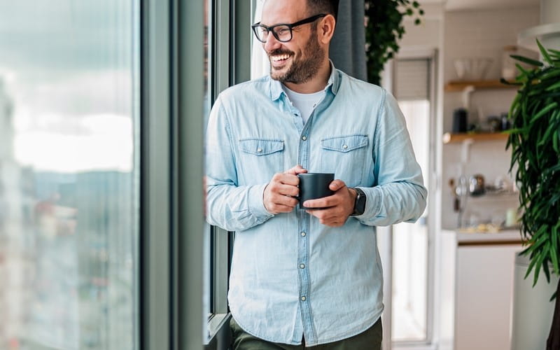 Light-skinned man holding a mug looks happily out a window